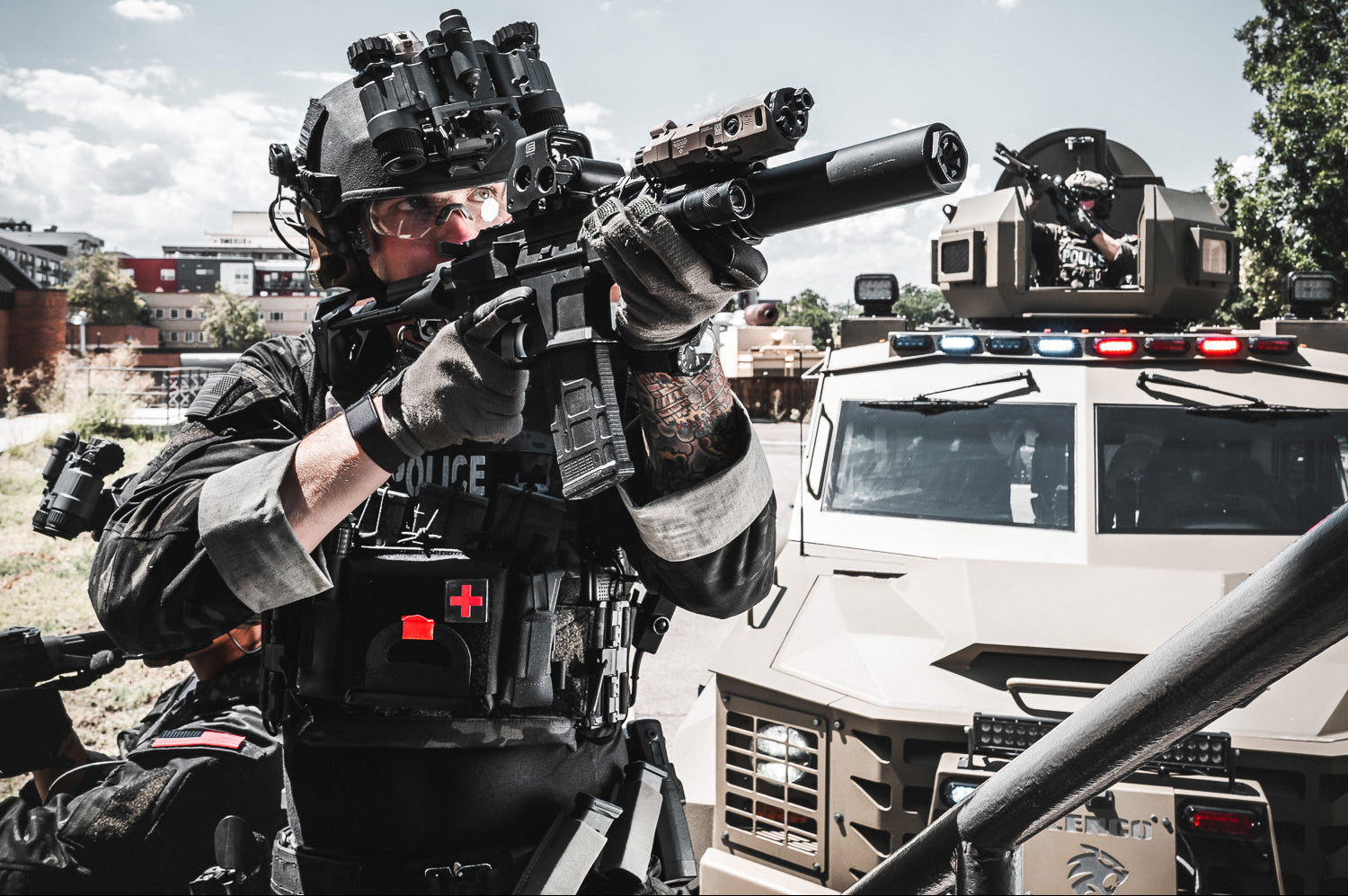 Person in tactical gear holding a weapon with a vehicle and buildings in the background